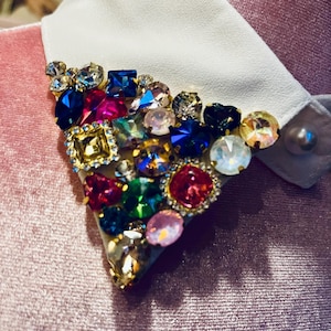 May include: A vibrant brooch, adorned with an array of colourful gemstones in shades of blue, red, and green, is attached to a white collar. The brooch sits on a pink velvet fabric, with a pearl button in view.