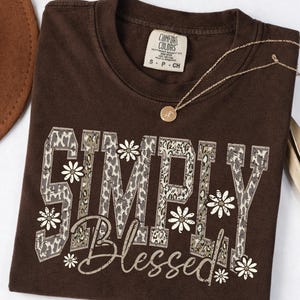 May include: A brown t-shirt with the words "SIMPLY BLESSED" in a leopard print design, accented with white daisy graphics. A gold necklace with a pendant is draped on the shirt. A brown hat is partially visible in the background.