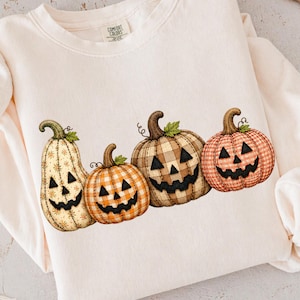May include: A cream-colored crewneck sweatshirt featuring a graphic of four jack-o'-lanterns with distinct patterns, including plaid and floral designs. The text DTF TRANSFERS is printed at the top. The garment has long sleeves and a ribbed collar.
