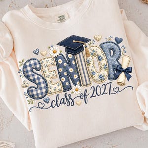May include: A cream-colored crewneck sweatshirt featuring a graphic print that reads SENIOR class of 2027. The letters are decorated with blue gingham, floral patterns, and daisies. A graduation cap sits on top, and a diploma with a blue bow is included.