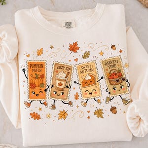 May include: A cream-colored crewneck sweatshirt featuring a graphic print of four anthropomorphic rectangular postage stamps. The stamps depict a pumpkin, a coffee cup, a slice of pie, and a basket of apples with the text Pumpkin Patch, Cozy Sip, Sweet Autumn, and Harvest Time.