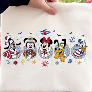 May include: Cream-colored tote bag featuring nautical-themed Disney characters: Goofy, Mickey Mouse, Minnie Mouse, Pluto, and Donald Duck. Each character is in a porthole, surrounded by nautical elements like anchors, seagulls, and starfish.