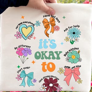 May include: Cream-colored tote bag with the words "it's OKAY to" in a retro font, surrounded by colorful illustrations and phrases like "Have bad days" and "Ask for help." The design includes flowers, bows, and stars.
