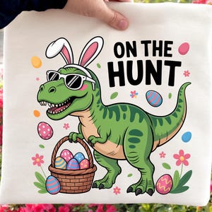 May include: A cream-colored tote bag featuring a green dinosaur wearing sunglasses and bunny ears, holding a basket of Easter eggs. The text "ON THE HUNT" is displayed in black. Easter-themed design.