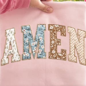 May include: A light pink pillow with the word "AMEN" embroidered in a collegiate style. The letters are made of fabric with floral and patterned designs, including white, blue, and brown. The pillow is held up by a person's hand.