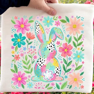May include: A square, off-white tote bag featuring a colorful Easter bunny design. The bunny is surrounded by pink, blue, and orange flowers and green leaves. The bunny has black spots and gold accents.