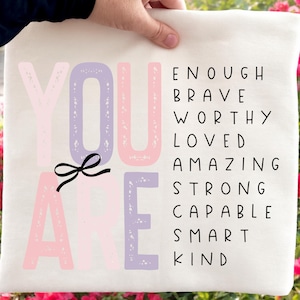 May include: A cream-colored pillow with the words "YOU ARE" in pink and lavender, with a black bow graphic. The right side of the pillow lists the words: ENOUGH, BRAVE, WORTHY, LOVED, AMAZING, STRONG, CAPABLE, SMART, KIND.