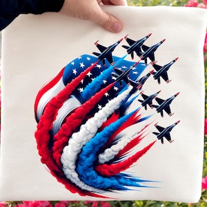 May include: A cream-colored tote bag featuring a patriotic design. The design includes an American flag with red, white, and blue smoke trails, and a formation of black fighter jets with red accents. The jets are flying in formation.