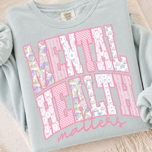 May include: A light blue sweatshirt with the words "MENTAL HEALTH matters" in a pink and floral design. The sweatshirt is folded on a neutral-colored surface.