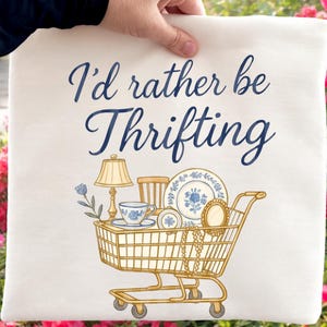 May include: Cream-colored tote bag with the text "I'd rather be Thrifting" in navy blue script. The bag features a shopping cart illustration filled with vintage items like a lamp, teacup, and plates. The cart is gold-colored.