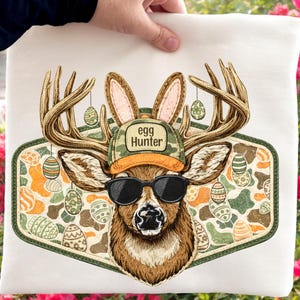 May include: A white tote bag featuring a deer wearing sunglasses, a camouflage hat with the text "egg Hunter", and bunny ears. The deer has antlers with Easter eggs hanging from them. The background has a camouflage pattern with Easter eggs.