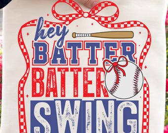 May include: A cream-colored baseball-themed pillow with the words "Hey Batter Batter Swing" in red and blue. A baseball bat and a baseball with a red bow are also featured in the design. The pillow is held up by a person.