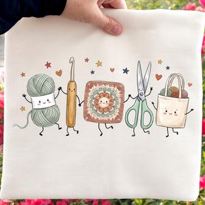 May include: A white tote bag featuring a graphic illustration of five anthropomorphic craft items holding hands: a ball of sage green yarn, a wooden crochet hook, a floral granny square, a pair of mint green scissors, and a beige tote bag.