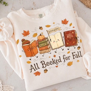 May include: A cream-colored crewneck sweatshirt featuring a graphic of three anthropomorphic books with faces, a pumpkin, and autumn leaves. The text reads "BOOKWORM" on a flag and "All Booked for Fall" below the books, with "COZY NOTES" on one cover.