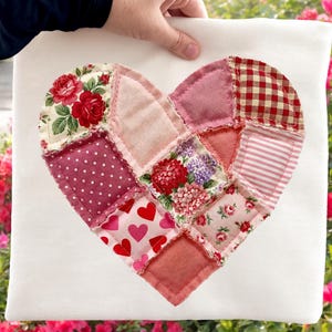 May include: A white square pillow featuring a patchwork heart design. The heart is composed of various fabric patterns, including floral prints, polka dots, and gingham in shades of red, pink, and white. The pillow is held by a person.
