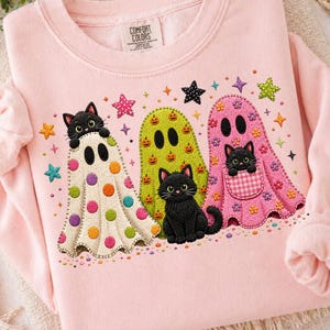 May include: A light pink crewneck sweatshirt with a Comfort Colors label at the neck. The graphic features three ghost figures: one cream with colorful polka dots, one lime green with pumpkin prints, and one pink with floral and gingham pocket details.