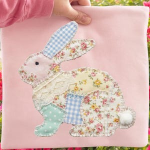 May include: A pink pillow with a patchwork bunny applique. The bunny features floral, gingham, and polka dot fabrics in pastel colors. A white pom-pom tail completes the design. The pillow is held up by a person.