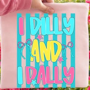 May include: A light pink pillow with the text "I Dilly and I Rally" in a colorful design. The text is in pink, yellow, and blue, with star and bow accents. The pillow is held up by a person.