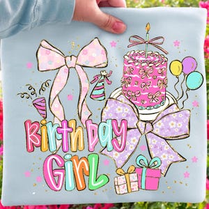 May include: A light blue t-shirt with a colorful "Birthday Girl" graphic. The design includes a pink cake with a candle, bows, confetti, balloons, and gift boxes. The graphic has a glittery gold outline.