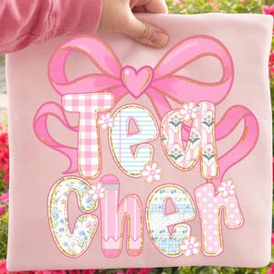 May include: A pink pillow with the word "Teacher" in a decorative font. The letters are adorned with patterns like gingham, stripes, and polka dots, and accented with small flower illustrations. A large pink bow and heart shape complete the design.