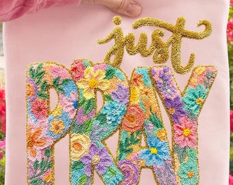 Könnte beinhalten: Ein hellrosa quadratisches Kissen mit den goldgestickten Worten "just PRAY" und bunten Blumenmustern. Das Kissen wird von einer Hand gehalten, mit einem Hintergrund aus rosa Blumen.
