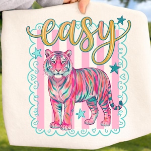 May include: A white tote bag featuring a colorful tiger illustration with pink, teal, and orange stripes. The word "easy" is written in a cursive font above the tiger, with stars and a scalloped border.