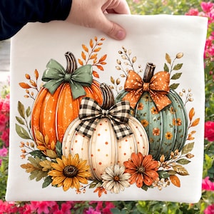 May include: A white fabric item featuring a graphic print of three pumpkins. One orange pumpkin has a sage green bow, a central white pumpkin with orange dots has a black and white checkered bow, and a teal pumpkin has an orange floral bow.
