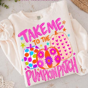 May include: A cream-colored crewneck sweatshirt featuring a graphic print with the text "TAKE ME TO THE PUMPKIN PATCH" in bright pink. The design includes three pumpkins decorated with colorful polka dots, pink daisies, and small pink hearts.