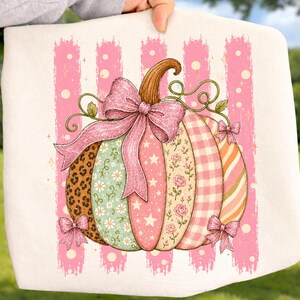 May include: A white fabric item featuring a graphic of a pumpkin with vertical segments in leopard print, floral patterns, stars, gingham, and stripes. The pumpkin is decorated with a large glittery pink bow and smaller bows at the base.