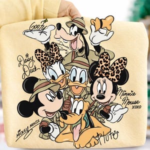 Puede incluir: Un bolso tote amarillo claro con personajes de Disney con atuendos de safari. Mickey Mouse, Minnie Mouse, Donald Duck, Daisy Duck, Goofy y Pluto est&aacute;n representados. Los nombres de los personajes y "XOXO" tambi&eacute;n est&aacute;n presentes.