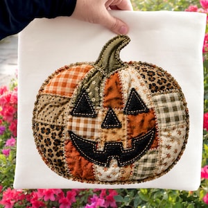 May include: A white t-shirt featuring a large, central pumpkin applique made from various fabric patches. The pumpkin is constructed with orange, brown, and cream textiles in plaid, floral, and leopard patterns, finished with black fabric eyes and a mouth.