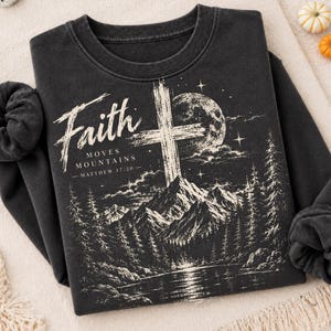 May include: A charcoal gray crewneck sweatshirt featuring a white graphic print of a cross, a full moon, and a mountain landscape with pine trees. The text reads Faith Moves Mountains Matthew 17:20 in a stylized white font.