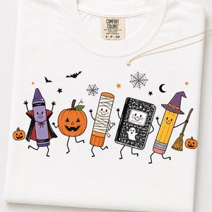 May include: A white t-shirt featuring a graphic print of five anthropomorphic school supplies dressed for Halloween. The design includes a purple crayon vampire, a jack-o'-lantern, a glue stick mummy, a composition notebook ghost, and a pencil witch.