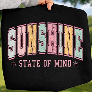 May include: Black tote bag with the word "SUNSHINE" in large, colorful, retro-style lettering. Below, the text "STATE OF MIND" is displayed in a smaller font. The bag is held up against a green background.