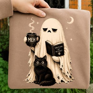May include: A tan sweatshirt featuring a graphic of a ghost holding a black mug labeled MEH and reading a book titled HOW TO CARE BUT NOT REALLY, with a black cat sitting in front of the ghost, all under a crescent moon and small stars.