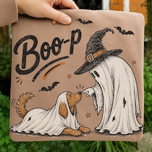 May include: A tan sweatshirt featuring a graphic print of a ghost wearing a witch hat touching the nose of a dog covered in a white sheet. The text "Boo-P" is printed in black above the figures, accompanied by small orange stars and black bats.