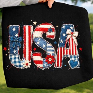 May include: Black fabric with the word "USA" in red, white, and blue patchwork letters. The letters are decorated with stars, bows, and flowers, evoking a patriotic theme.