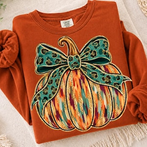May include: A burnt orange long-sleeve sweatshirt with a Comfort Colors label at the neck. The front features a large graphic of a pumpkin with vertical stripes in orange, teal, and gold, topped with a large teal leopard print bow.