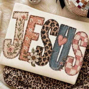 May include: A cream-colored sweatshirt featuring the word JESUS in large, block-style letters. Each letter has a unique pattern: floral, pink daisy, leopard print, denim with a pink heart, and pink gingham check, all with a stitched-look border.