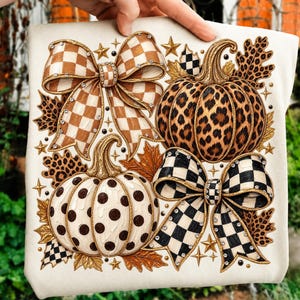 May include: A cream-colored t-shirt featuring a graphic print of two pumpkins, one with a leopard print and one with brown polka dots, accented by two large checkered bows, autumn leaves, and small gold stars with embedded clear rhinestones.