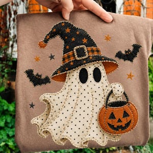 May include: A tan t-shirt featuring a central applique of a cream-colored ghost with black polka dots. The ghost wears a black witch hat with orange stars and a plaid band, holding an orange jack-o'-lantern bucket, surrounded by black bats.