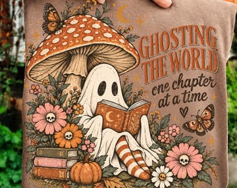 Könnte beinhalten: Ein braunes T-Shirt mit einer Grafik eines Geistes, der unter einem großen Pilz ein Buch liest, umgeben von Blumen mit Totenkopf-Mitten, Schmetterlingen und einem Bücherstapel. Der Text lautet GHOSTING THE WORLD one chapter at a time.