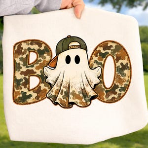 May include: A white fabric item featuring a graphic print of the word BOO in a brown, green, and tan camouflage pattern. A white ghost character wearing a green baseball cap is positioned in the center, replacing the letter O in the word.