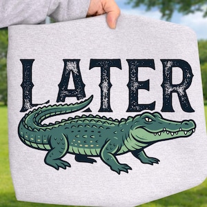 May include: A light gray tote bag featuring a green alligator illustration with the word "LATER" in distressed black lettering. The alligator is detailed with various shades of green and white accents.