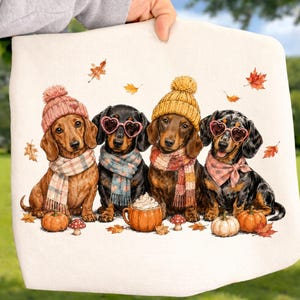 May include: A white fabric square featuring an illustration of four dachshunds wearing winter accessories. The dogs wear knit hats, scarves, and heart-shaped glasses, surrounded by autumn leaves, small mushrooms, and a pumpkin mug with whipped cream.