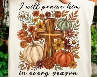 May include: A cream-colored t-shirt featuring a graphic of a wooden cross surrounded by pumpkins and autumn flowers. The text reads I will praise him in every season and Psalm 146:2. The design includes white, orange, yellow, and green colors.