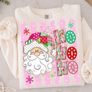 May include: A cream-colored crewneck sweatshirt featuring a graphic of a Santa Claus face with a patchwork hat. The design includes the text HO HO HO in colorful, patterned letters, surrounded by pink vertical brushstrokes and snowflakes.