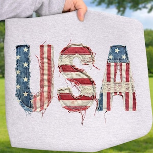 May include: A light gray tote bag with the word "USA" appliqued on the front. The letters are made from denim and fabric with a distressed American flag design, featuring red and white stripes and blue with white stars.