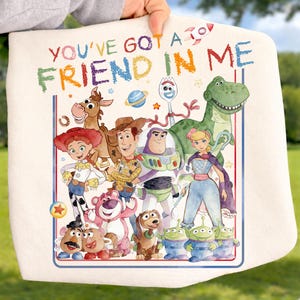May include: A white fabric item featuring a colorful graphic of Toy Story characters including Woody, Buzz Lightyear, Jessie, Rex, and others. The text "YOU'VE GOT A FRIEND IN ME" is printed in multicolored, crayon-style lettering above the characters.