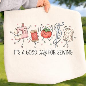 May include: A cream-colored tote bag featuring a colorful graphic of a pink sewing machine, a red thread spool, a tomato pincushion, a silver needle, and a floral fabric square with arms and legs. The text reads, IT'S A GOOD DAY FOR SEWING.